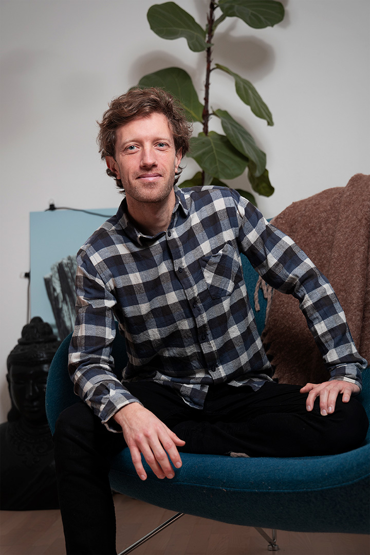 Psykolog i København psykolog Morten Skyum Nielsen sidder i rum til terapi i en ternet skjorte skorte og sorte bukser i en blå sofa med det ene ben oppe i sofaen og kigger ind i kameraet
