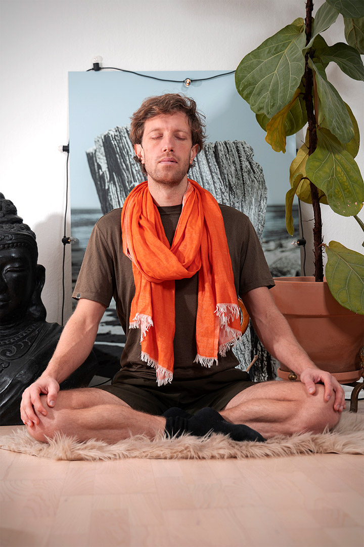 Psykolog i København psykolog Morten Skyum Nielsen sidder i forbindelse med en session med terapi i mindfulness og spiritualitet og dyrker yoga i t-shirt og shorts med orange tørklæde om halsen i skrædderstilling med lukkede øjne foran en sort statue af Buddha, et stort billede af pæle og en høj grøn plante i en lerkrukke
