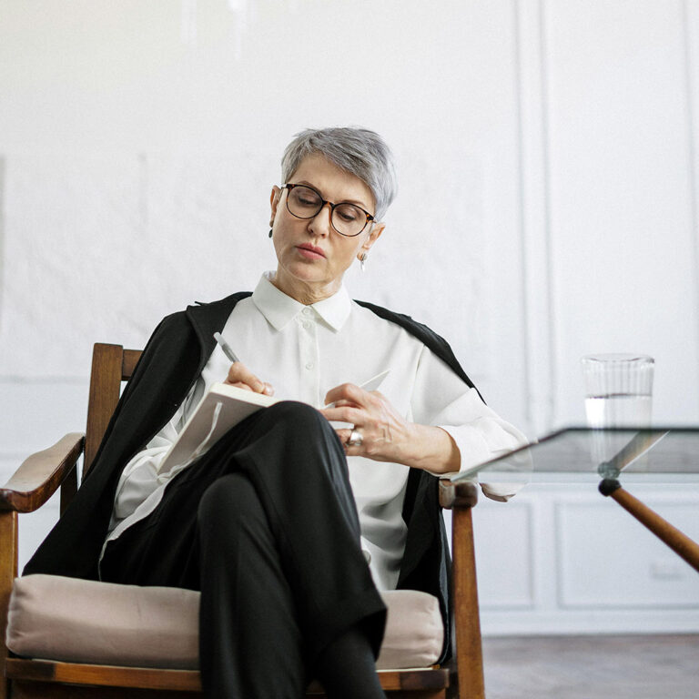 Moden gråhåret kvinde med kort hår og sorte briller sidder med benene over kors i en hvid skjorte med sorte bukser i en stol hos Psykolog i København psykolog Morten Skyum Nielsen og modtager ledersparring